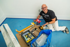 A professional painter kneels on a protected floor, smiling while organizing painting tools and supplies for an interior painting project.